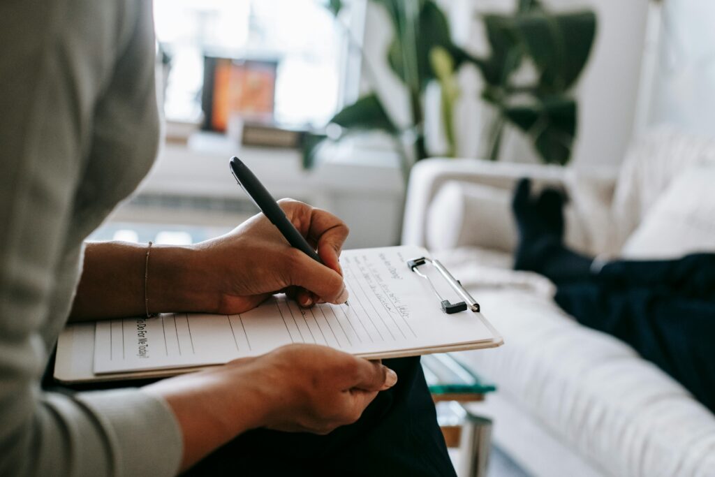 A person writes notes on a clipboard with a pen. In the background, someone relaxes on a sofa, feet up. The scene is calm and focused.