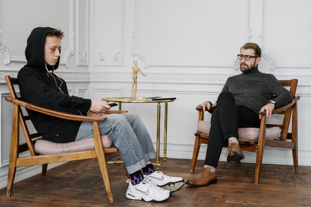 Two people sit facing each other in a minimalist room. The person on the left, wearing a hoodie and headphones, looks down at their phone. The person on the right, in glasses and a sweater, sits cross-legged and appears attentive. A small table with a wooden figure and notebook is between them. The mood is contemplative.