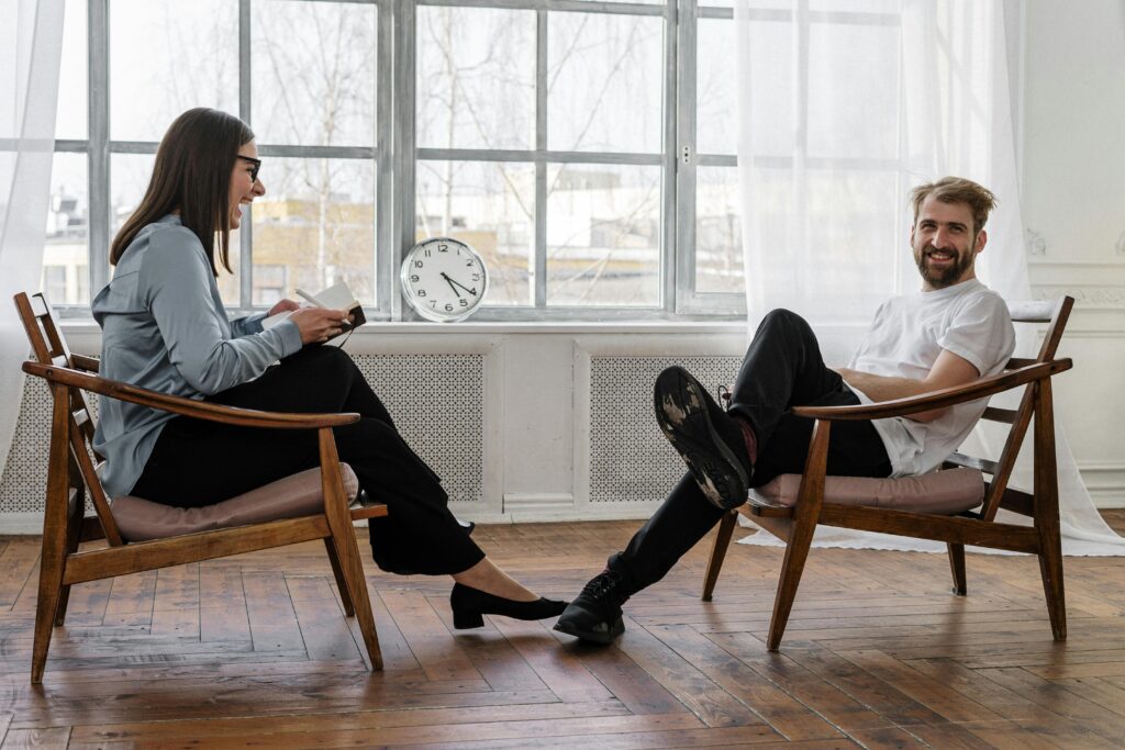 A woman and a man are sitting opposite each other in wooden chairs, smiling and engaged. The woman is writing in a notebook. A clock shows 10:10.