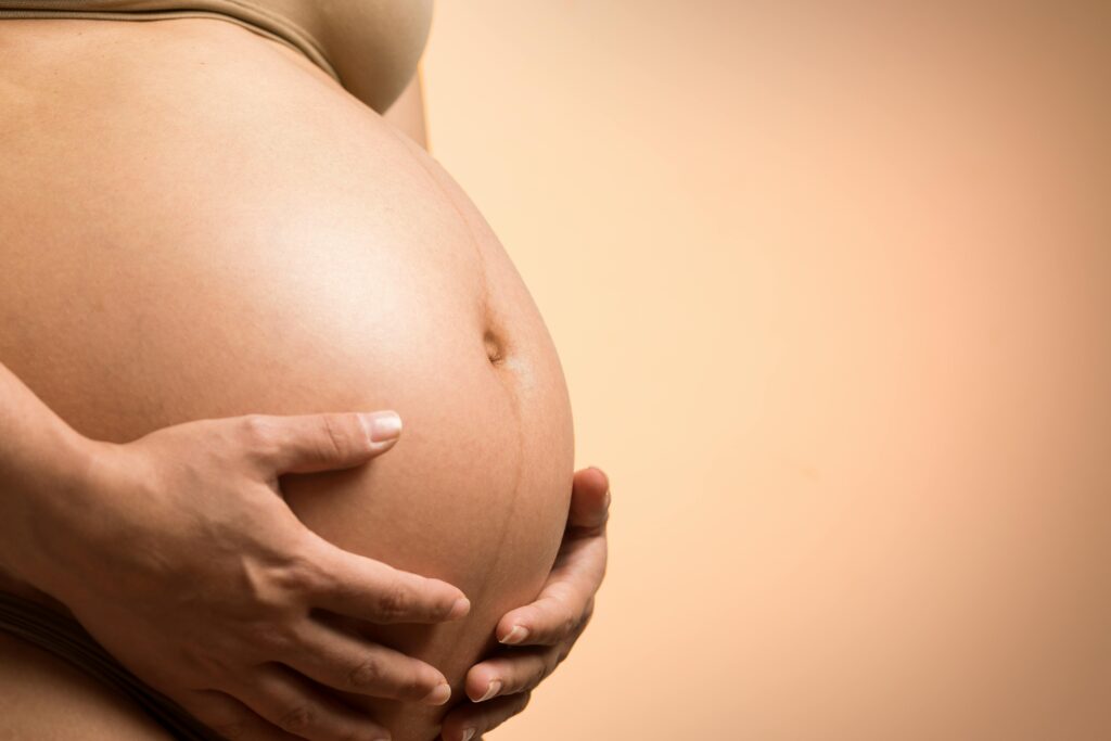Close-up of a pregnant belly with hands gently cradling it against a soft beige background, conveying warmth, care, and anticipation.