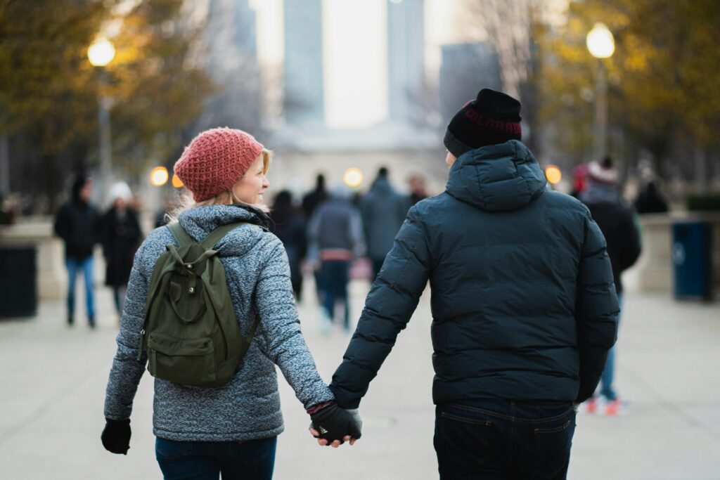 Couple holding hands and walking in a winter cityscape. Woman in a red beanie and gray jacket; man in a black coat. Warm ambiance, blurred crowd.