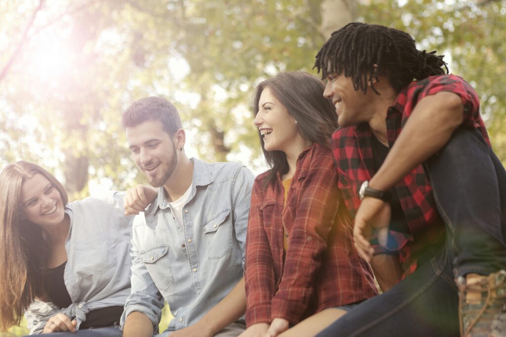 A group of four friends sitting outdoors, laughing and chatting under trees. Sunlight filters through the leaves, creating a warm and cheerful atmosphere.
