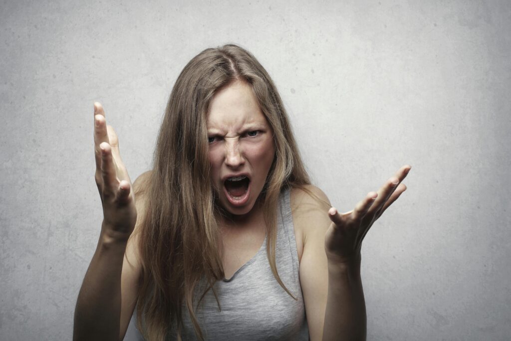 Woman with long hair angrily gesturing and shouting, wearing a gray tank top. Her expression conveys frustration. Gray background adds intensity.