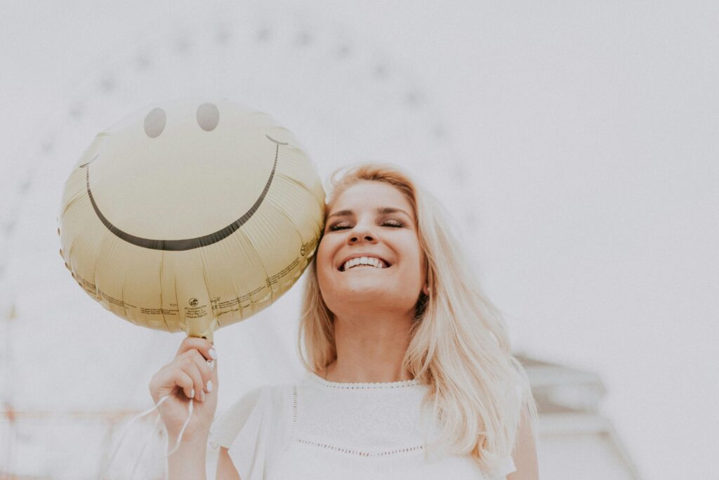 Woman joyfully holds a yellow smiley face balloon outdoors, eyes closed, with a big smile. Blurred Ferris wheel in background adds a playful feel.