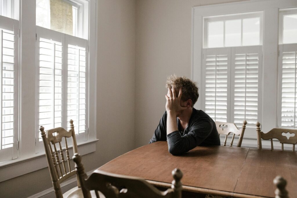 A person sitting at a wooden table in a bright room with large windows and shutters. Their face is resting in one hand, conveying a sense of contemplation.