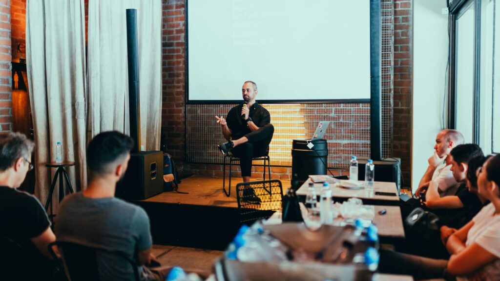 Speaker sits on stage with a microphone, engaging an audience in a modern loft with brick walls and a projector screen. Attentive and casual atmosphere.