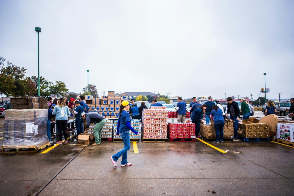 Group of volunteers in a parking lot organizing food donations. Stacks of boxes and canned goods are visible. The mood is collaborative and busy.
