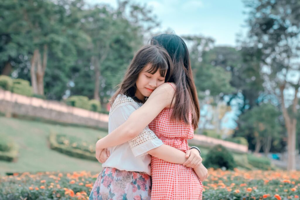 Two young women embrace warmly in a park, with one wearing a red checkered dress and the other a floral skirt. They appear content and serene, surrounded by greenery and flowers.