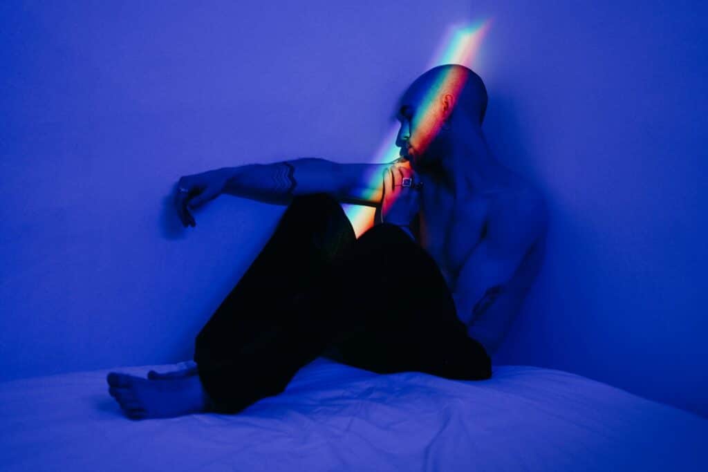 Shirtless person with a tattoo on their arm sits against a blue wall, illuminated by a colorful rainbow light. The scene conveys a contemplative mood.