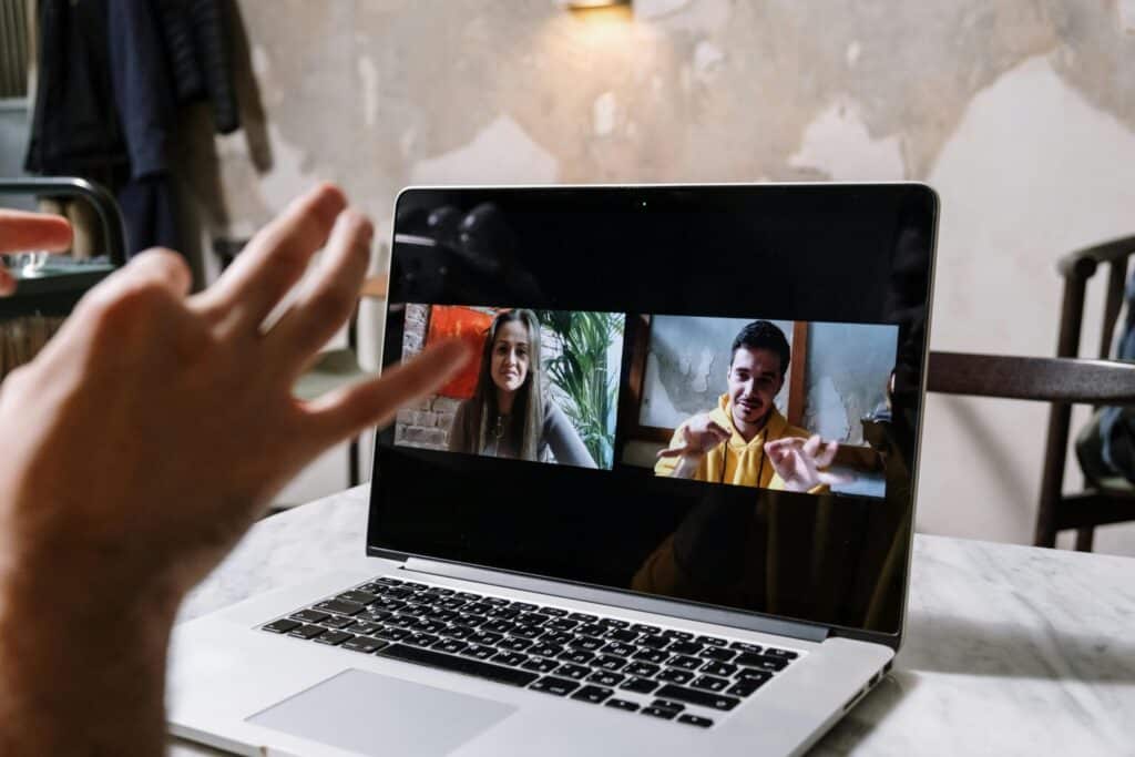 A laptop shows two people in a video call; a woman smiles on the left, while a man gestures on the right. The mood is friendly and engaging.