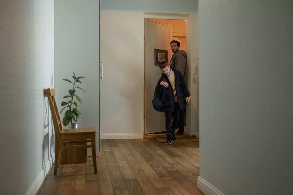 A young boy and a man enter a warmly lit room, the boy smiling with a bag, while the man follows. A chair with a potted plant is visible on the wooden floor.