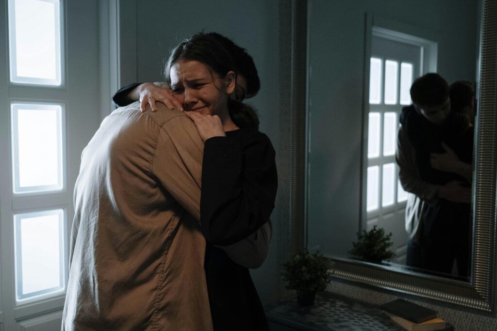 A woman hugs another person tightly, expressing distress while crying. They are in a dimly lit room, with their reflection visible in a mirror nearby.