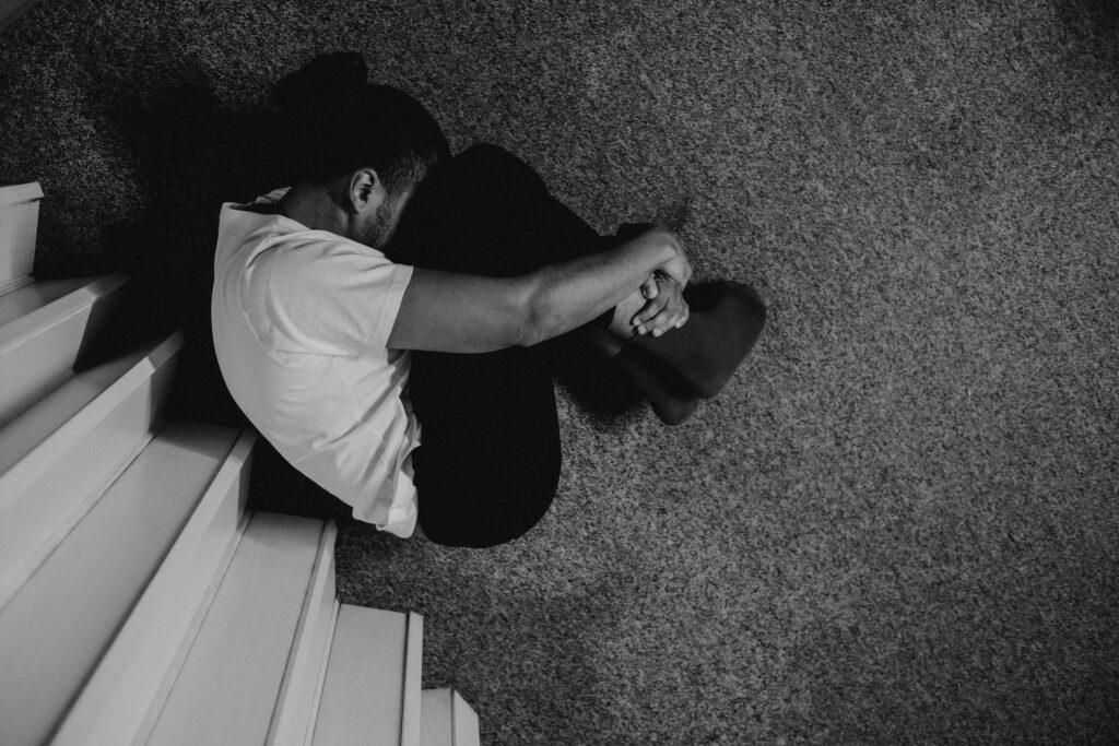 A person in a white shirt sits curled up on carpeted stairs, holding their knees, conveying a sense of introspection or melancholy in a dimly lit setting.