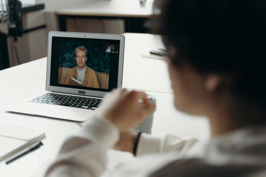 A person on a video call sits at a white desk, looking at a laptop screen showing another person smiling and holding a book, conveying a relaxed atmosphere.