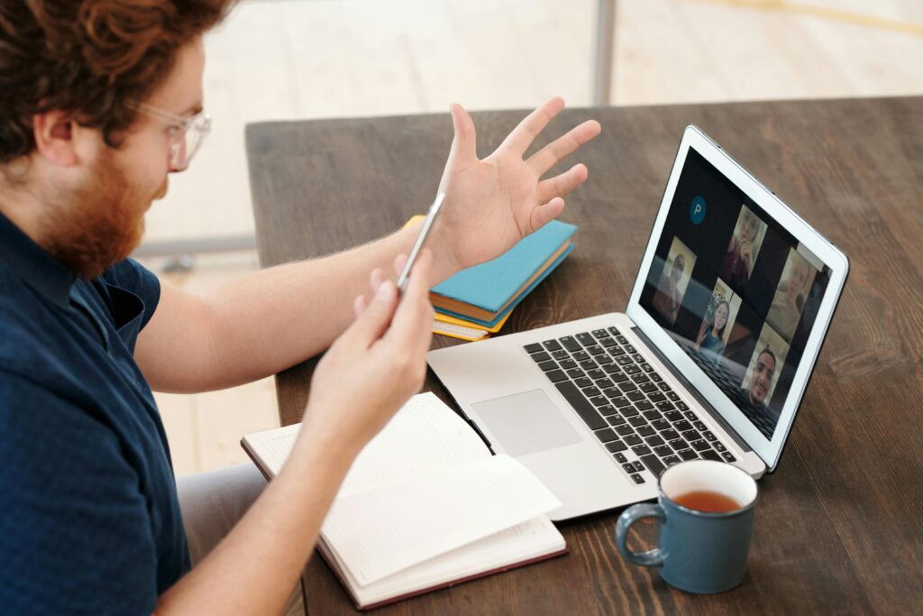 A man with wavy hair, glasses, and a blue shirt gestures with his hand during a video call on a laptop. An open notebook and tea cup sit nearby.
