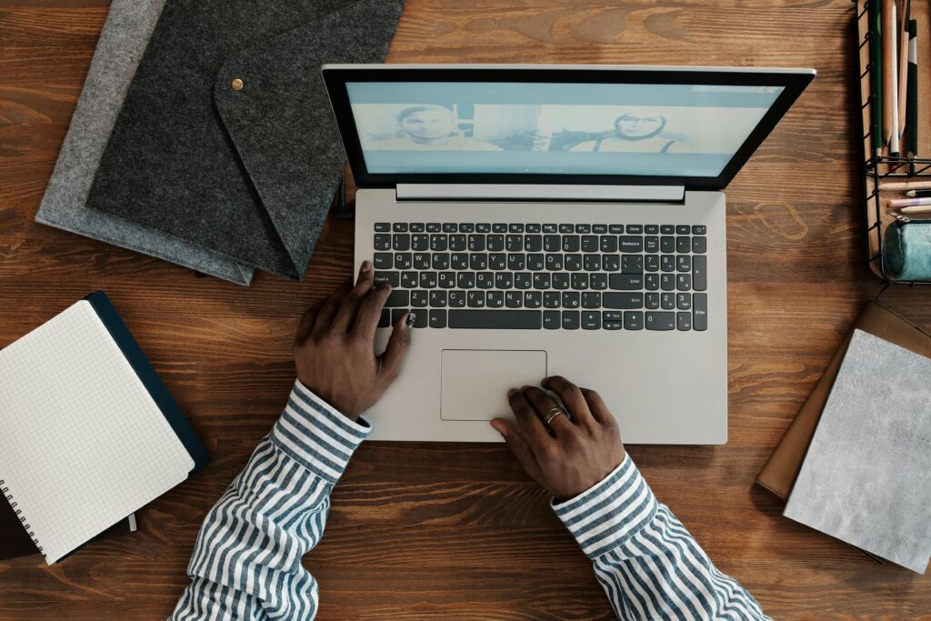 A person wearing a striped shirt types on a laptop showing a video call. Surrounding items include notebooks, folders, and pens, suggesting a work setting.