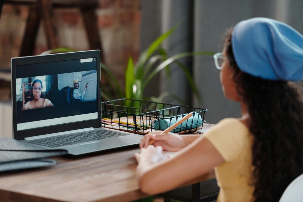 Girl in yellow shirt and blue beret sits at a wooden desk, watching a laptop screen showing a virtual meeting with two people, one holding a whiteboard.