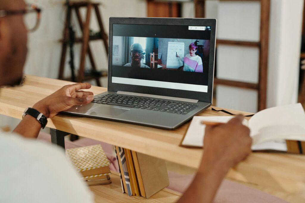 A person participates in a virtual meeting on a laptop, viewing two people. One points to a whiteboard. Open notebook and pen are near, showing focus.