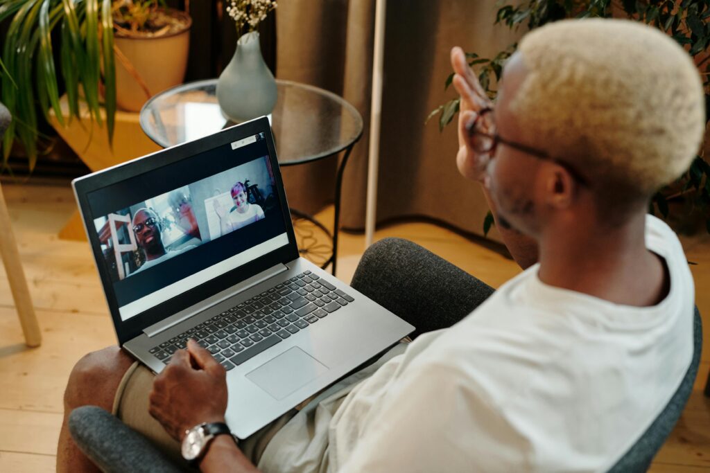 Person with short blonde hair participates in a casual video call on a laptop, sitting in a cozy room with plants. The mood is relaxed and social.