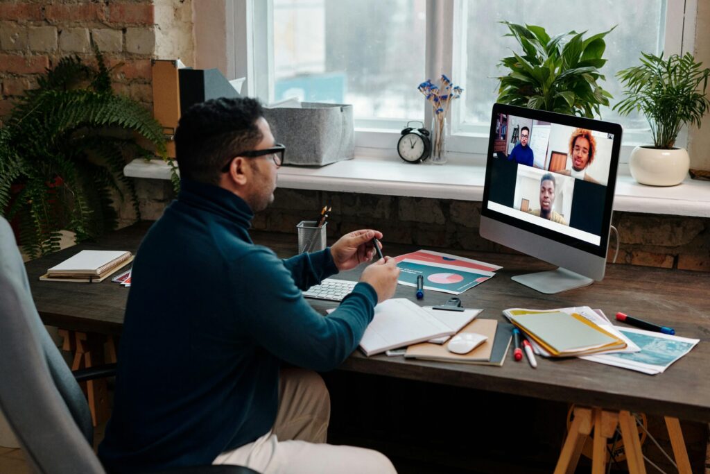 Man in a blue sweater sitting at a desk engaged in a video call with four people on a computer screen. Desk has notebooks and plants nearby. Cozy atmosphere.