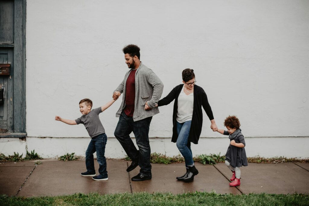 A joyful family of four walks hand-in-hand on a sidewalk. The parents are in the middle, holding hands with their smiling children on each side.