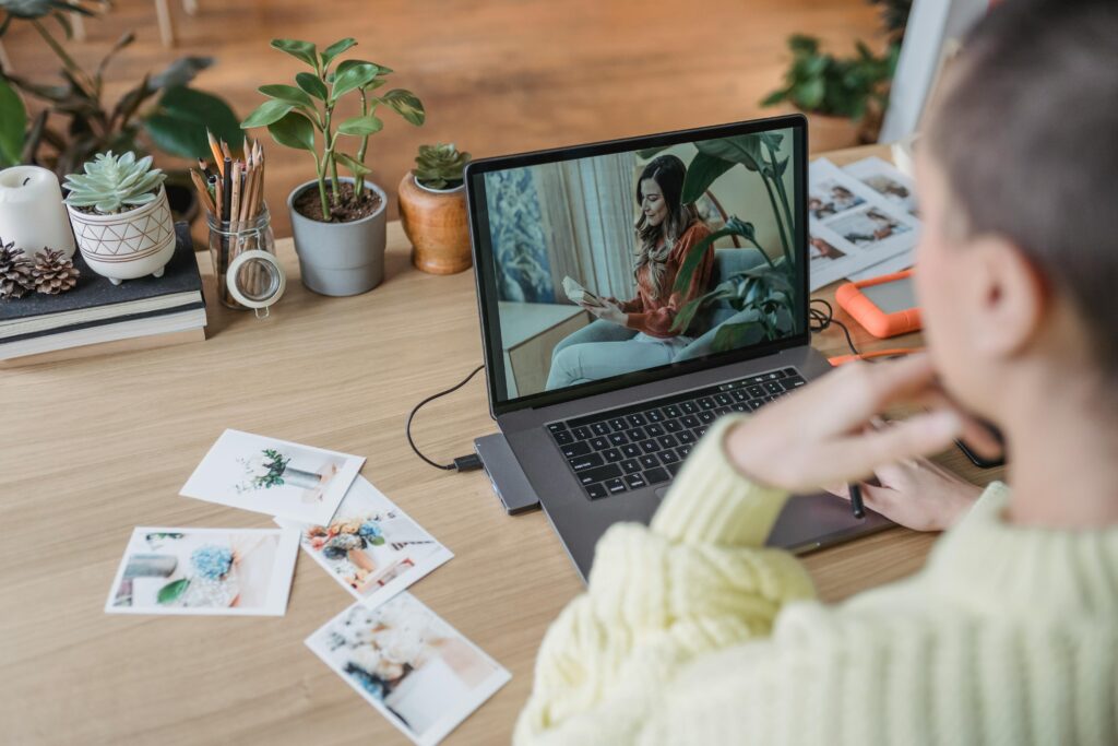 A person in a cozy sweater watches a video call on a laptop at a wooden desk with houseplants and scattered photos, creating a warm, creative workspace.