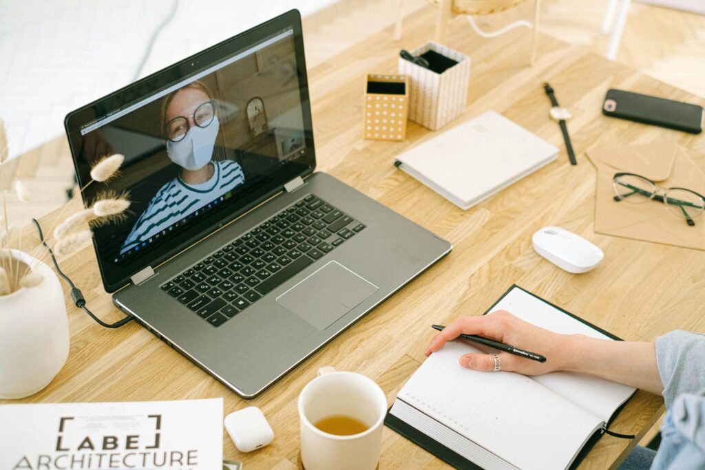 A person on a laptop video call, wearing glasses and a mask, sits at a wooden desk with notepad, mug, and stationery, creating a focused work setting.