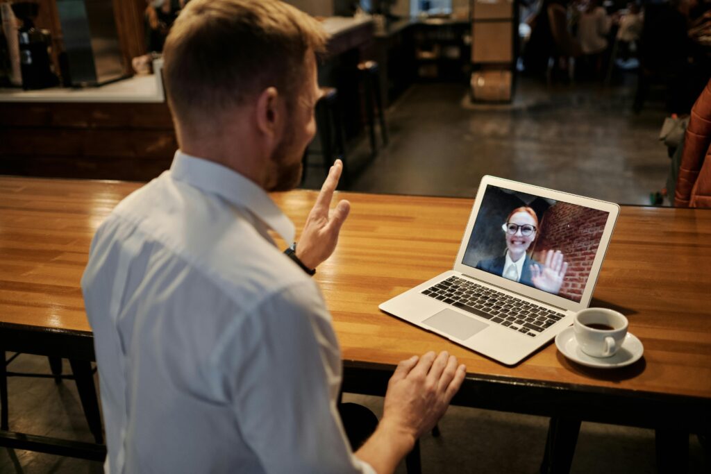 A man in a white shirt sits at a table, waving at a laptop screen. On the screen, a woman with glasses smiles and waves back. A cup of coffee is nearby.