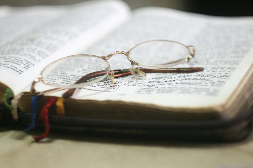 Open book with gold-frame glasses resting on it. Pages contain text with multicolored bookmarks visible, conveying a scholarly and reflective mood.