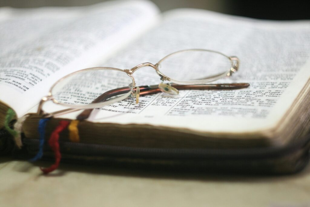 Open book with gold-frame glasses resting on it. Pages contain text with multicolored bookmarks visible, conveying a scholarly and reflective mood.