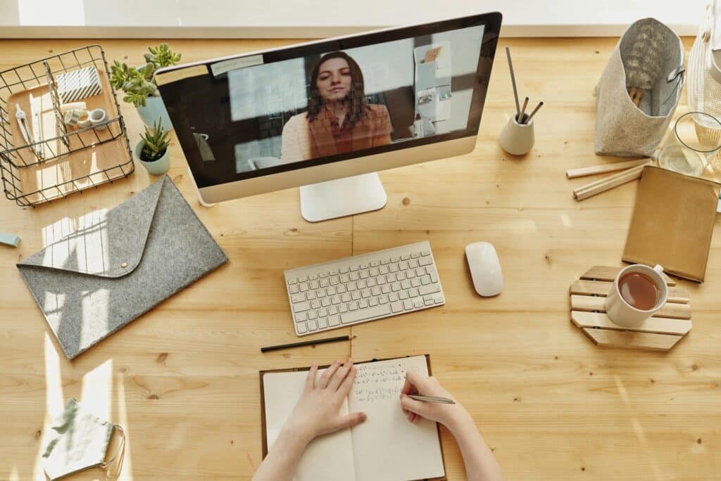 A person takes notes in a notebook at a wooden desk, facing a computer displaying a video call. The desk is organized with plants, stationery, and a cup of tea, conveying a productive atmosphere.