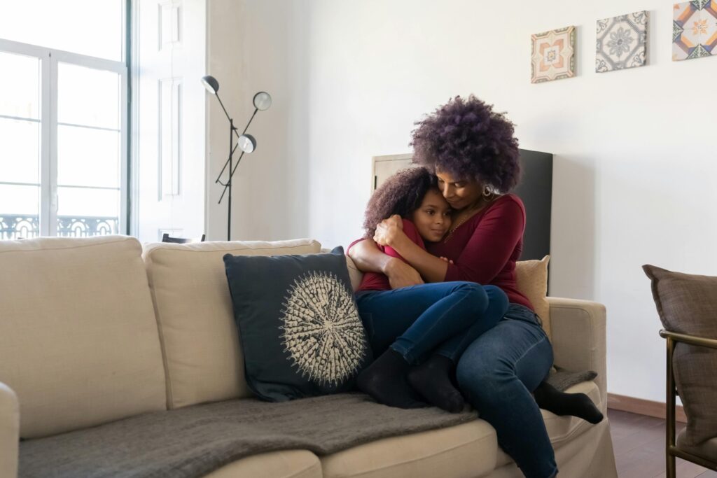A mother and daughter sit on a beige sofa, embracing warmly. The room is softly lit with cozy decor, creating a comforting and loving atmosphere.