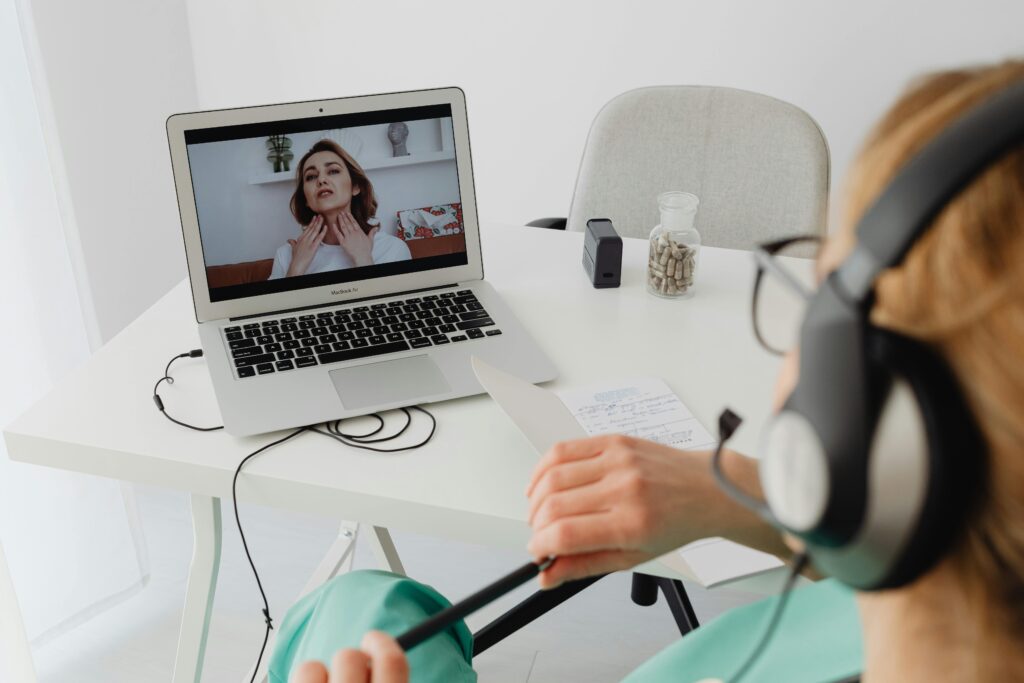 A medical professional in green scrubs and headphones takes notes during a telehealth video call. A patient gestures to her neck on the laptop screen.
