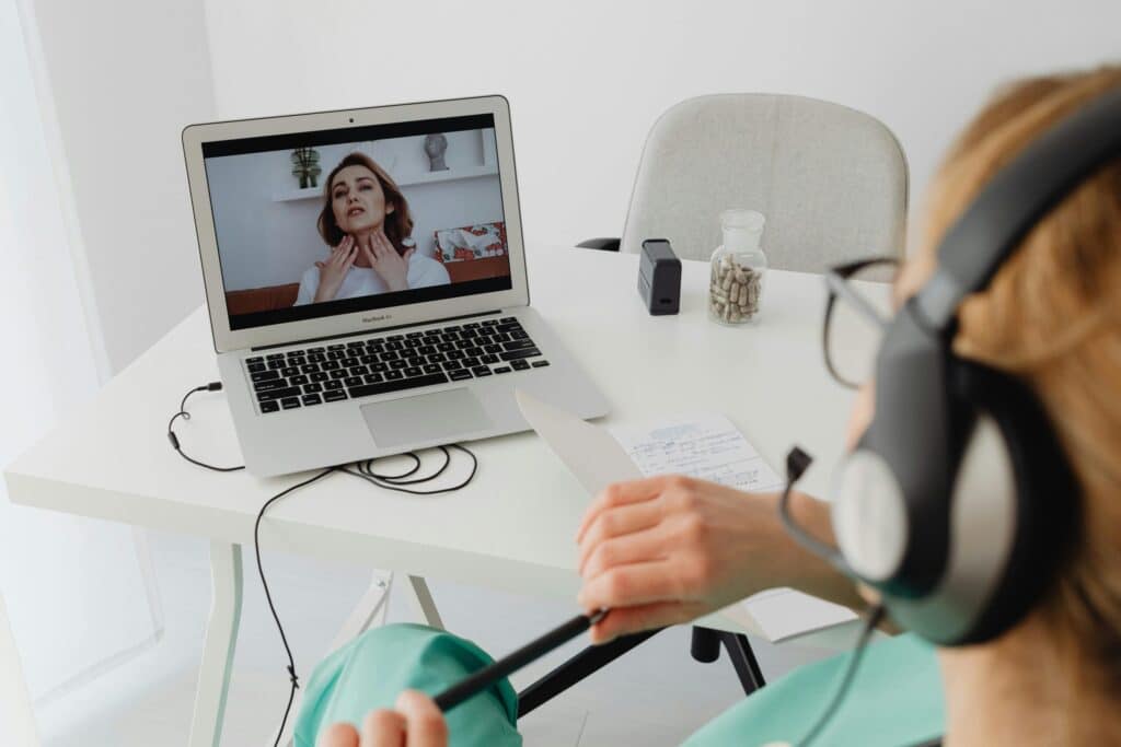 A medical professional in green scrubs and headphones takes notes during a telehealth video call. A patient gestures to her neck on the laptop screen.