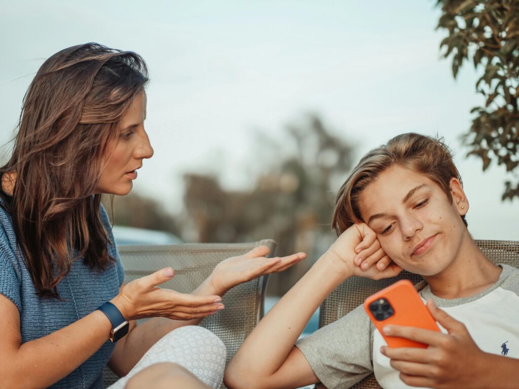 A woman talks expressively to a relaxed teenager, who smirks while looking at their phone. The setting is outdoors, suggesting a casual conversation.