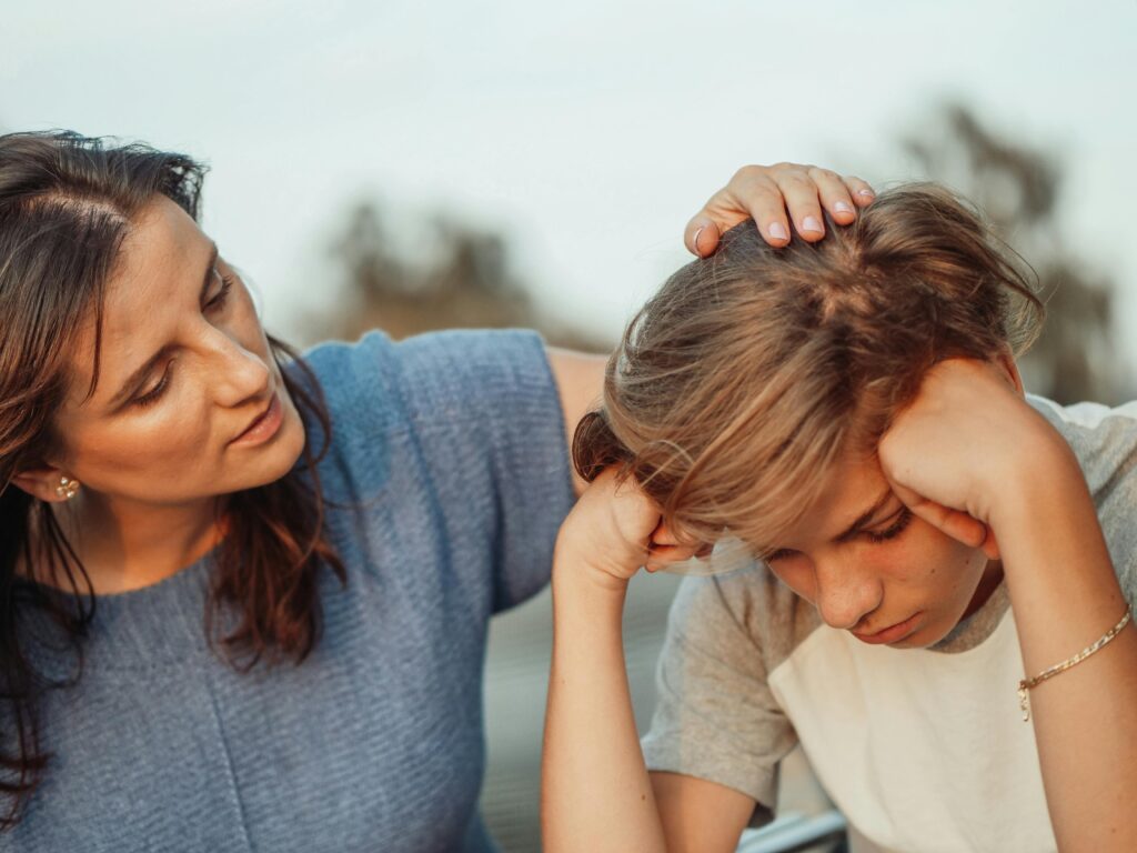 A woman in a blue shirt gently comforts a distressed child in a white and gray shirt by placing a hand on their head, conveying empathy and support.