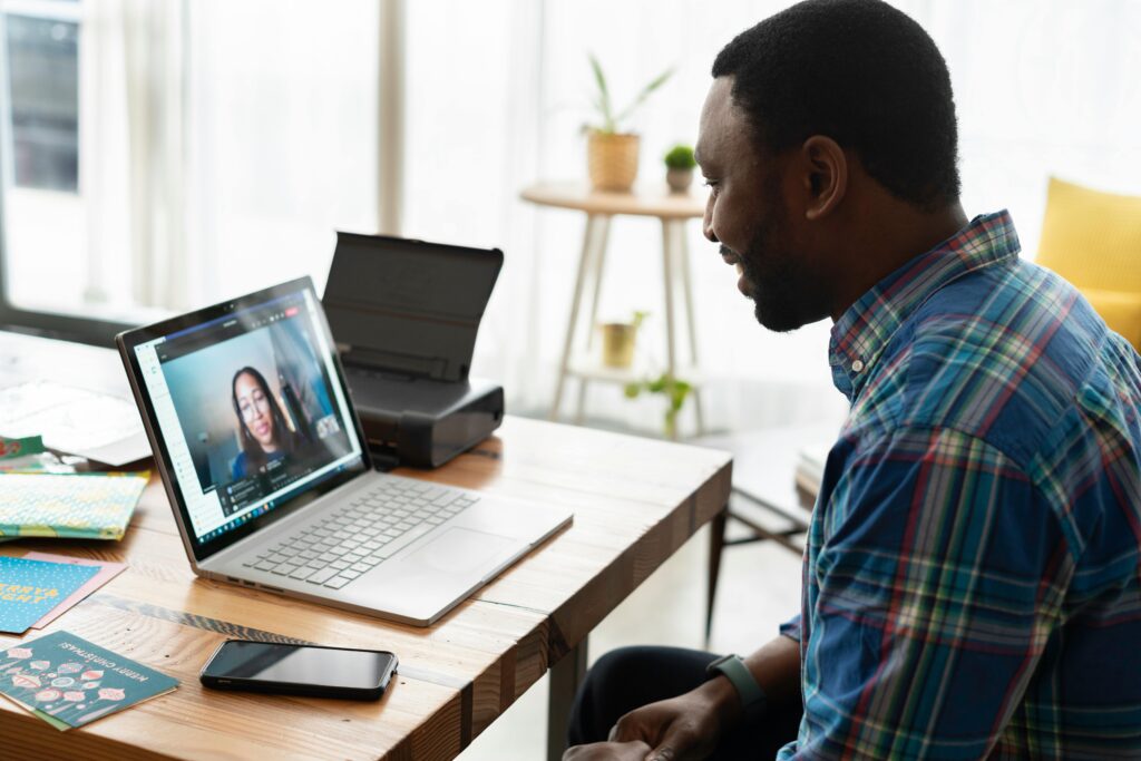 A man in a plaid shirt sits at a wooden desk, engaged in a video call on a laptop. The room is well-lit with a modern, casual atmosphere.