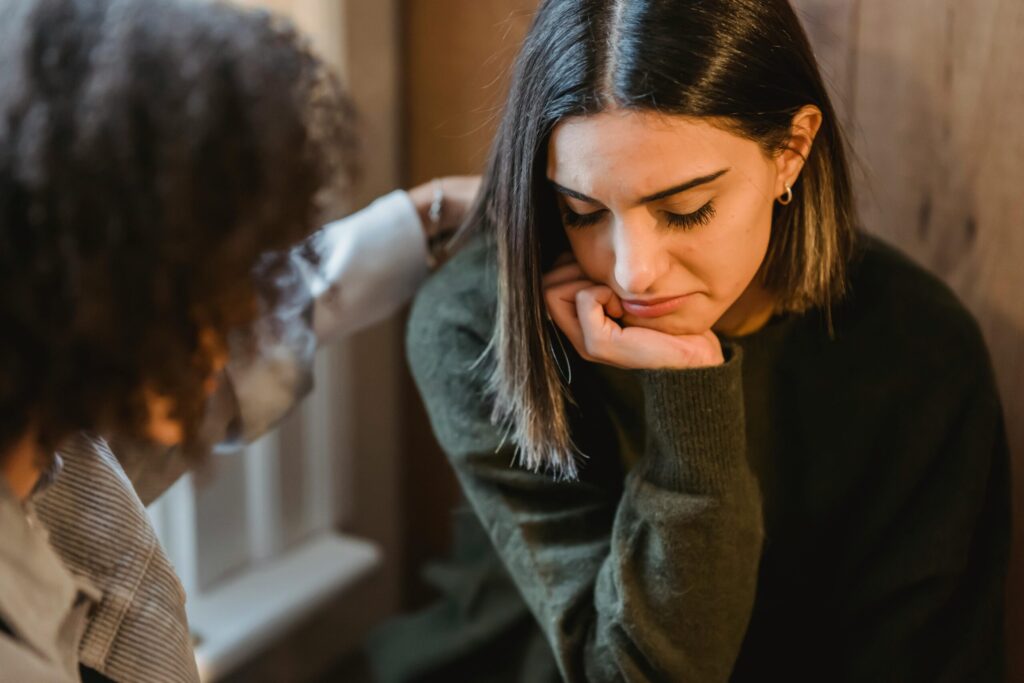 A woman with shoulder-length hair looks down, appearing sad, resting her chin on her hand. Another person, blurred, offers comfort by placing a hand on her shoulder.