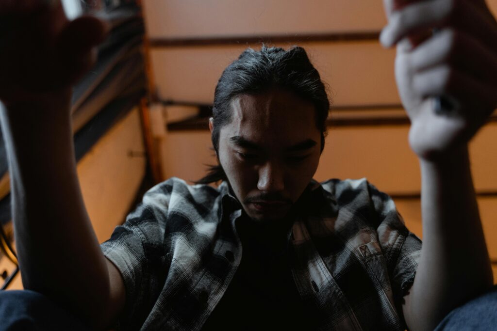 Man with long hair and a checkered shirt sits in dim light, looking down with a serious expression. Arms are raised, creating an introspective mood.