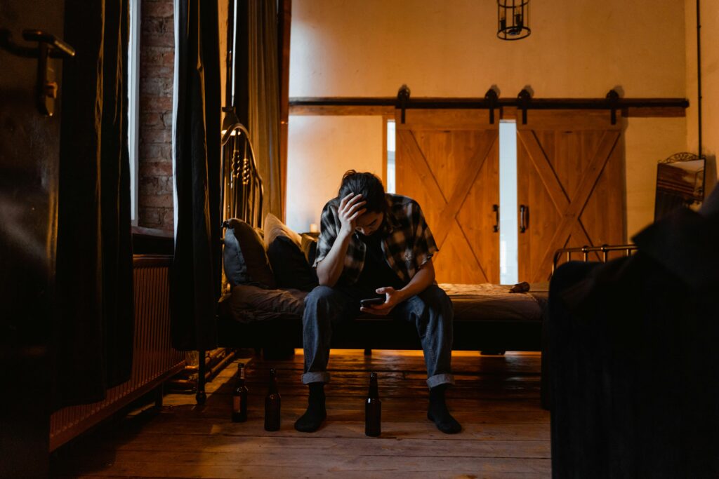 A person sits on a bed in a dimly lit room, hand on head, looking at a phone. Three empty beer bottles are on the floor, conveying a sense of sadness.