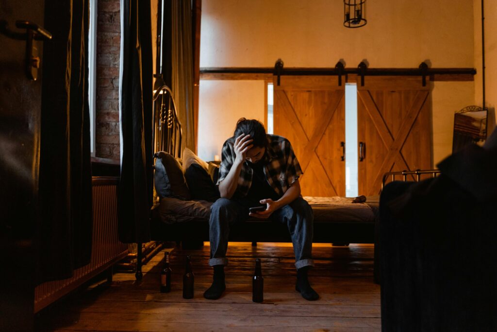 A person sits on a bed in a dimly lit room, hand on head, looking at a phone. Three empty beer bottles are on the floor, conveying a sense of sadness.