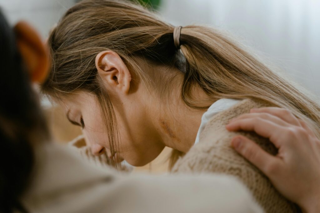 A woman with a brown ponytail leans forward, her face partially hidden, showing a bruise on her neck. A comforting hand rests on her shoulder.