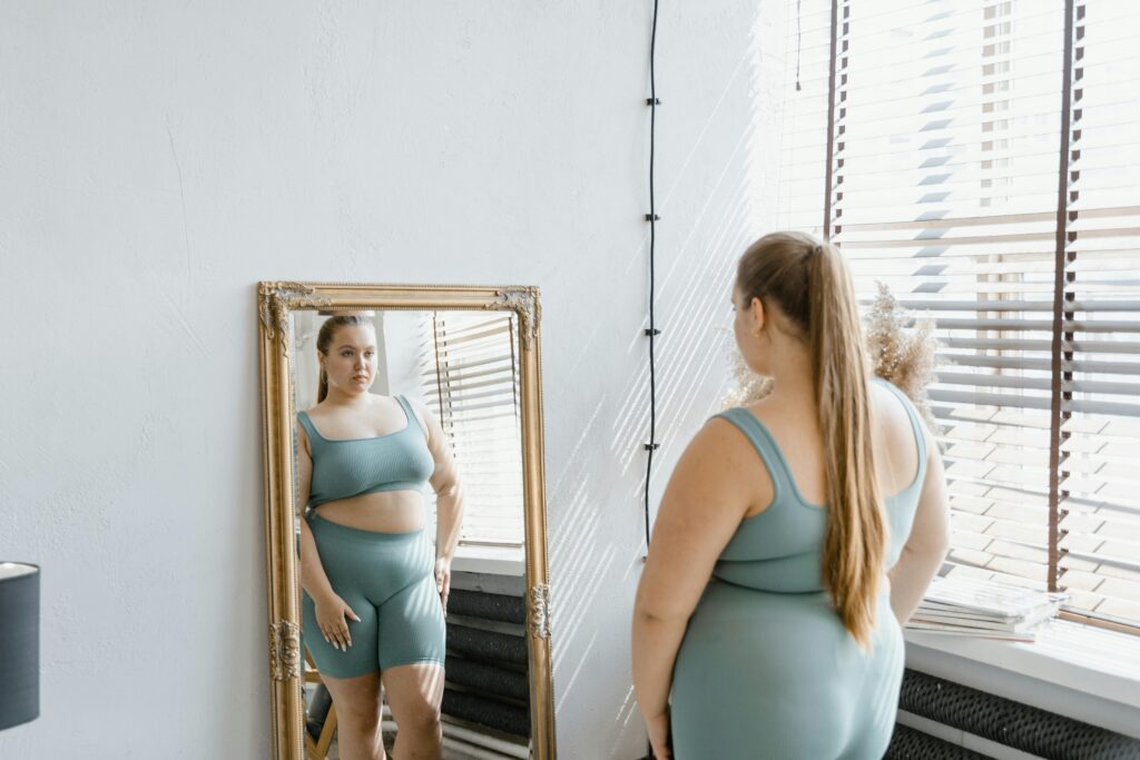 A woman in teal activewear gazes at herself in a full-length mirror. Sunlight streams through blinds, casting shadows, creating a contemplative mood.