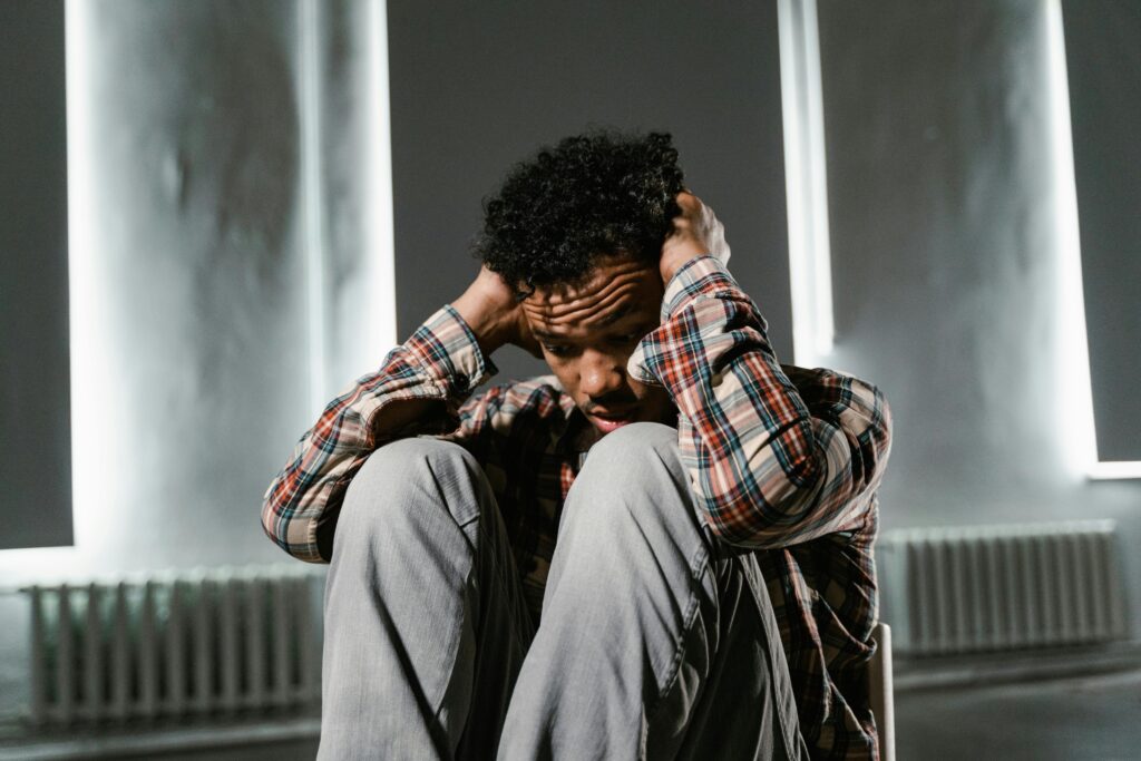 A man sits on a chair in a dimly lit room, holding his head with both hands, wearing a plaid shirt and jeans, appearing distressed and thoughtful.