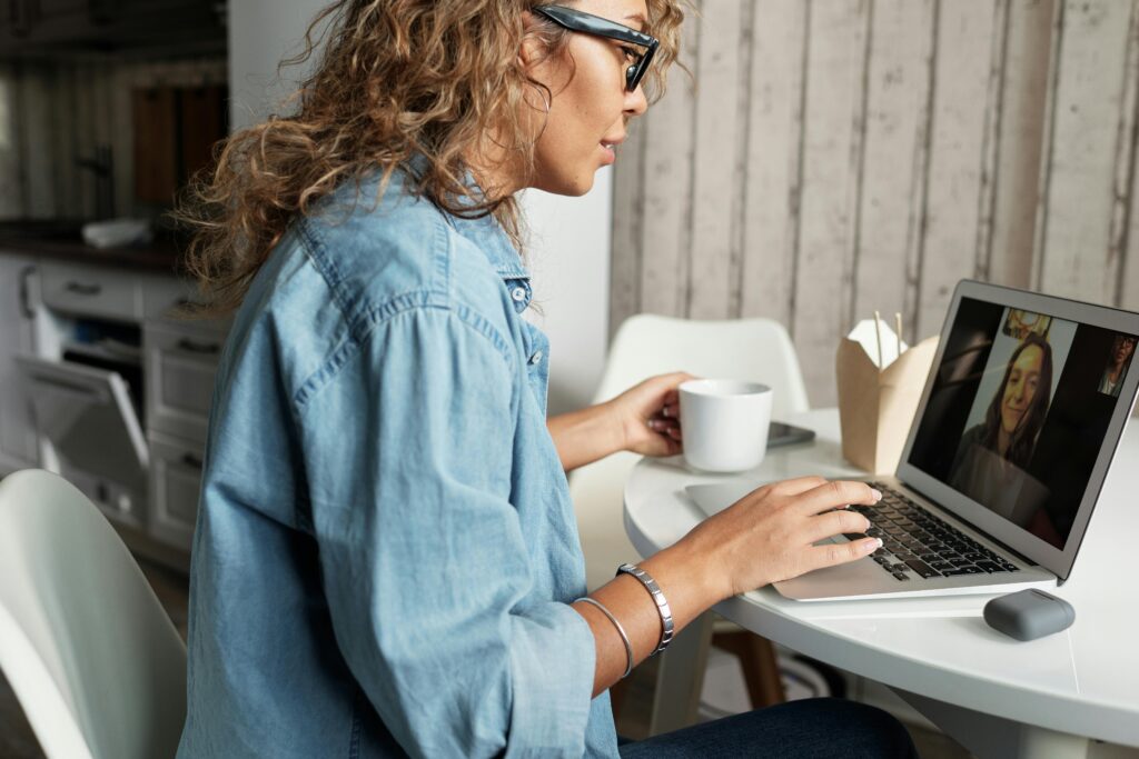 A woman in a denim shirt and glasses is having a video call on a laptop at a white table, holding a mug. The scene feels casual and focused.