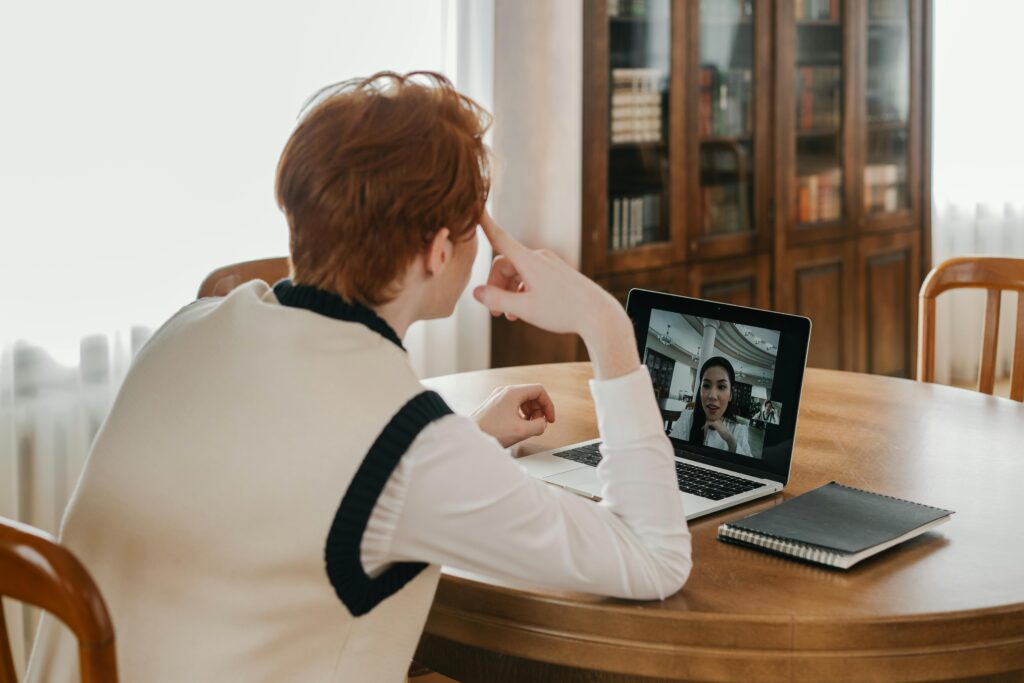 A person sits at a wooden table engaged in a video call on a laptop, with a notebook nearby. The room is light-filled, with bookshelves in the background.