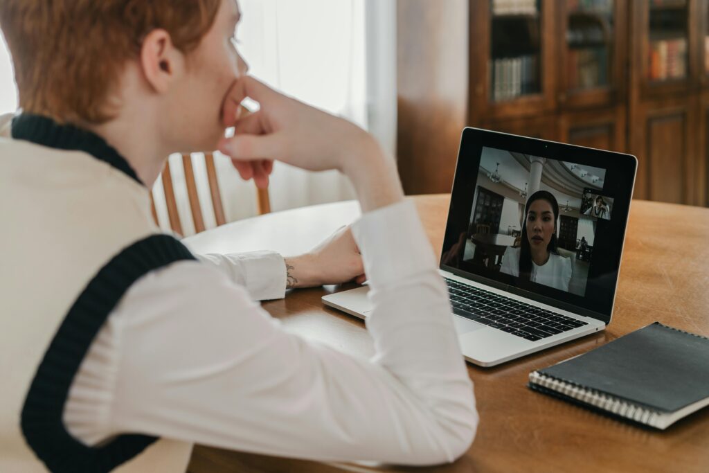 Person in a white shirt engaged in a video call on a laptop at a wooden table. Notebook beside them. Focused and attentive atmosphere.