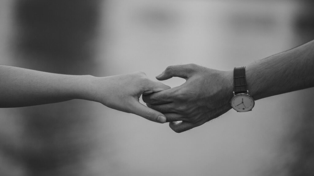 A black-and-white image showing two hands gently holding each other. One hand wears a watch, evoking a sense of connection and intimacy.