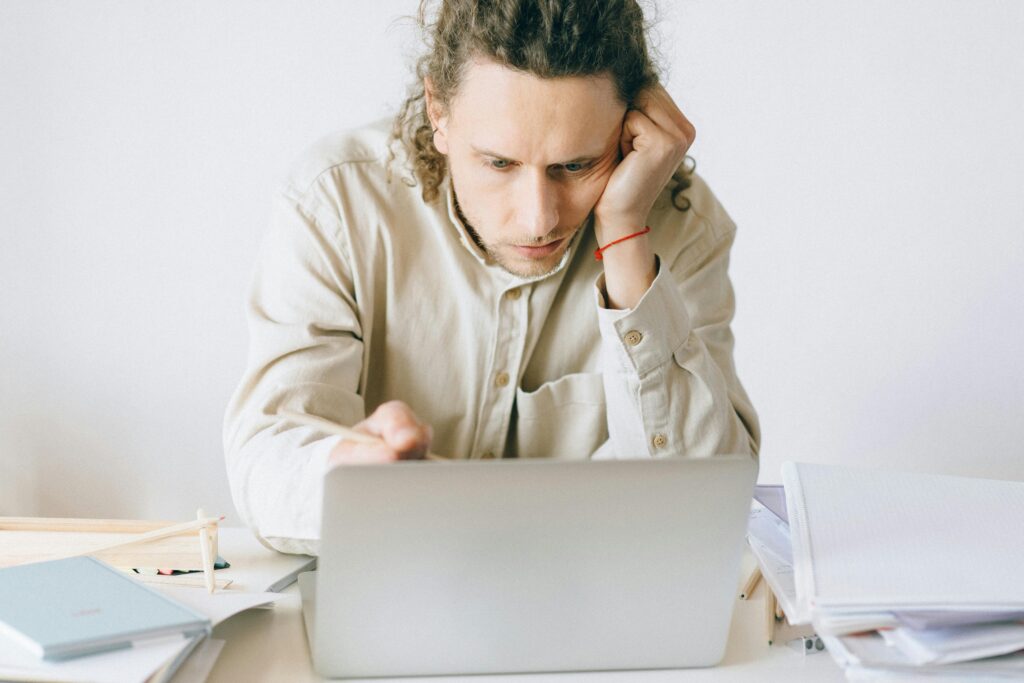 A man with curly hair, wearing a beige shirt, leans on his hand, concentrating intently on a laptop. Papers and notebooks clutter the desk.