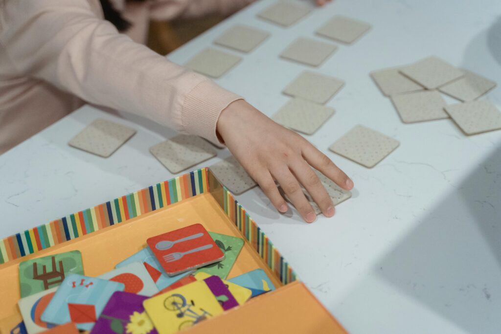 A child's hand reaches for a square card on a table, playing a memory match game. Colorful cards with images lie in a striped box nearby.